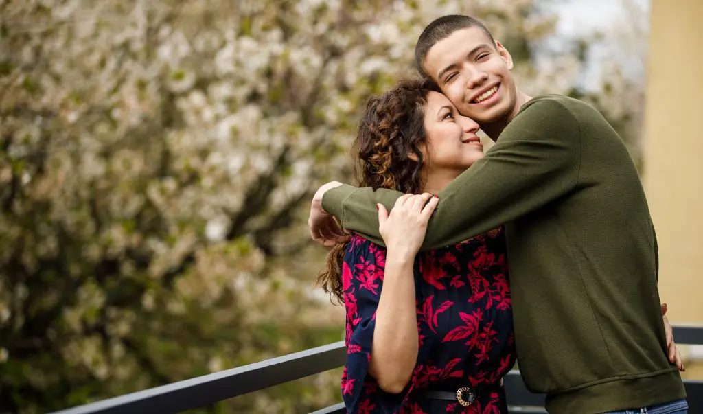A teenager embraces his mother while they pose for a picture outdoors.